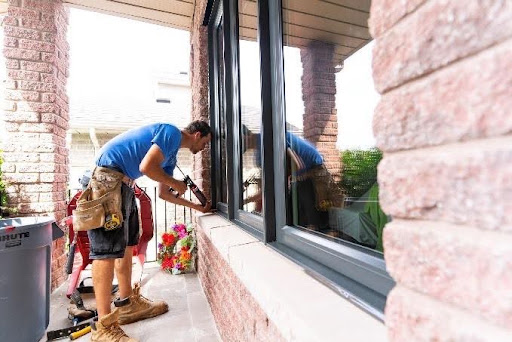 Professional window installer applying sealant around a modern black exterior window frame on a brick home.