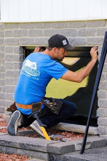 A technician in a blue shirt installing or repairing a basement window frame into a stone brick exterior wall.
