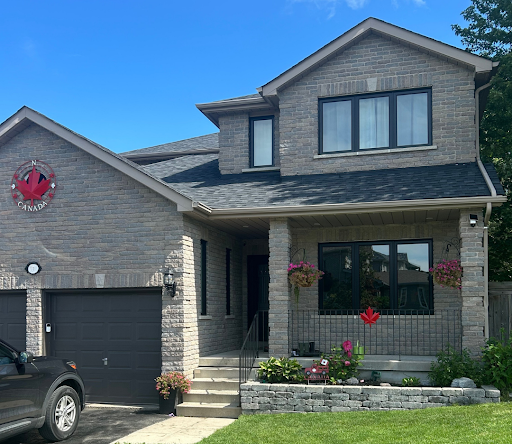 Exterior view of a two-story light stone house featuring black window frames, dark garage door, front porch with hanging flowers, and red Canadian maple leaf decorations.